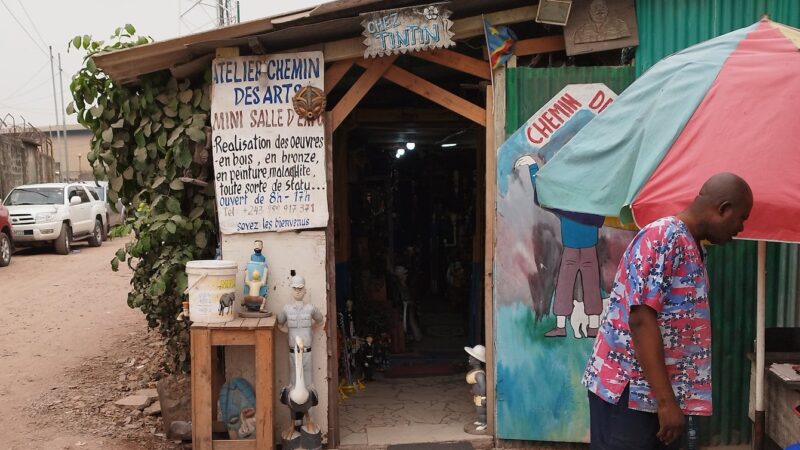 Auguy standing proudly in front of his atelier, surrounded by his sculptures and artworks, showcasing his dedication to preserving Congo's cultural heritage.